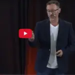 Speaker standing on a TED Talk stage delivering a presentation on the healing power of frequency, with a spotlight overhead and a minimalist backdrop.